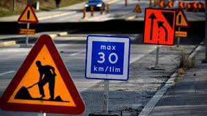 Orange vägarbetesskylt med symbol för vägunderhåll, omgiven av andra vägskyltar, inklusive fartbegränsning.