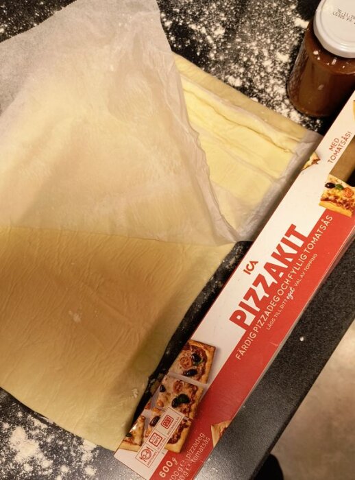 Förpackning för pizzakit med deg och tomatsås, delvis utrullad deg på bakplåtspapper, burk med tomatsås på en mjölat bänk.