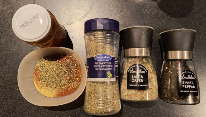 Kryddor för pizzabullar: pizzakrydda, salt och örter, svartpeppar och en skål med kryddblandning står på en mörk yta.