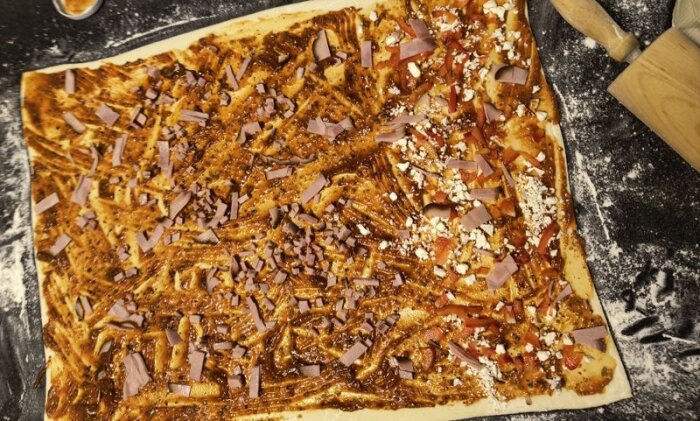 Rektangulär pizzadeg utrullad med kryddig tomatsås, skinka och ost, redo för att rullas till pizzabullar.