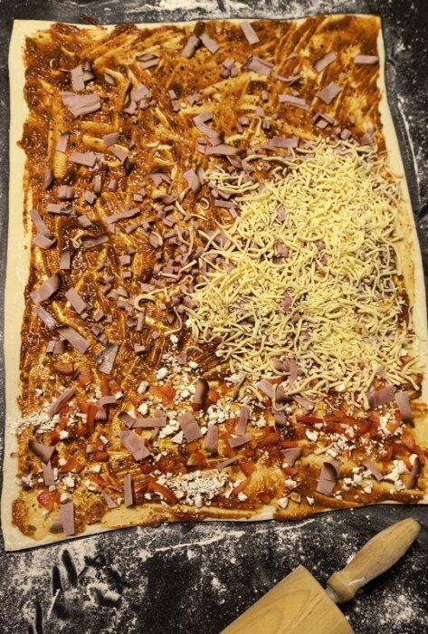 Rektangulär pizzadeg täckt med tomatsås, skinka, ost och kryddor, kavlad ut på mjölat bord bredvid kavel.