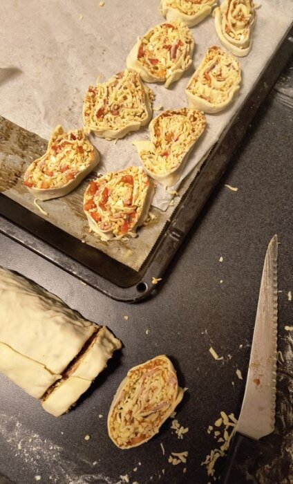 Pizzabullar på en bakplåt redo för gräddning, fyllda med skinka, ost, paprika och fetaost. En brödkniv och deg syns också på arbetsytan.