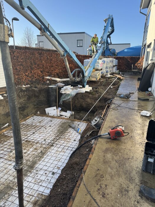 Bilden visar ett poolbygge med grävd grop, gjuten betongplatta och armering. En arbetare och utrustning syns vid poolen.