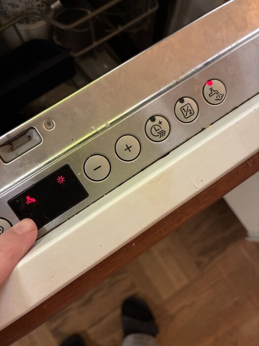 Diskmaskinspanel med knappar, display visar kransymbol och stjärna; rött ljus tänd. En finger pekar på kontrollpanelen.