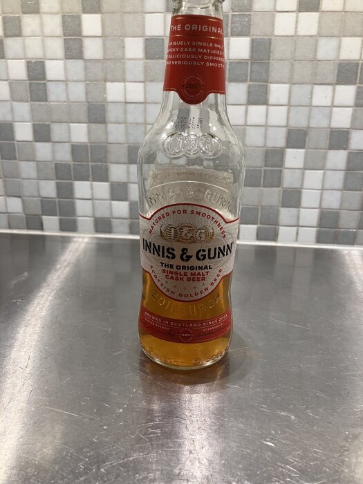 Tom flaska av Innis & Gunn original single malt cask beer på en rostfri yta med kaklad bakgrund.