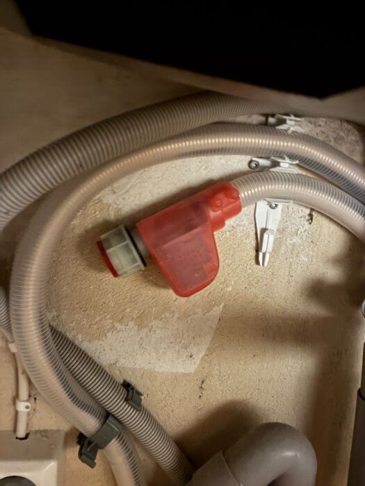 Gråa flexibla rör och en röd plastdel på en vägg, fastsatta med vita beslag.