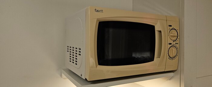 Gulnad mikrovågsugn med rundade hörn och frontpanel med två stora rattar placerad på en hylla.