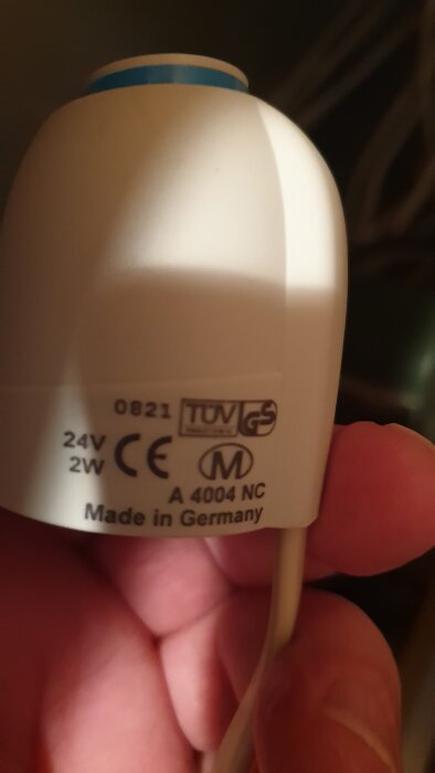 En vit komponent med etikett "Made in Germany" och flera certifieringssymboler, inklusive TUV och CE, hålls i en hand.