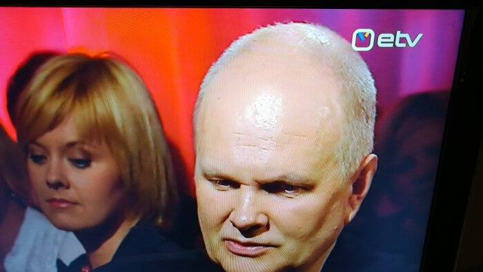 En närbild på två personer som tittar ned framför en röd bakgrund med logotypen för ETV i det övre högra hörnet.