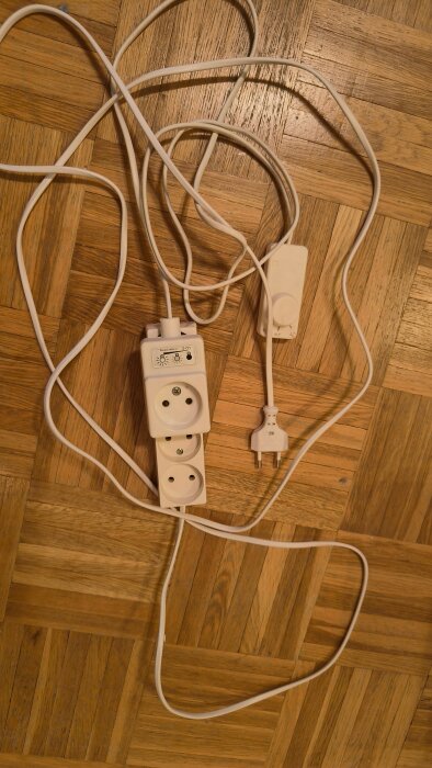 Vit kabel med två eluttag och en gammal dimmer ligger på ett parkettgolv.