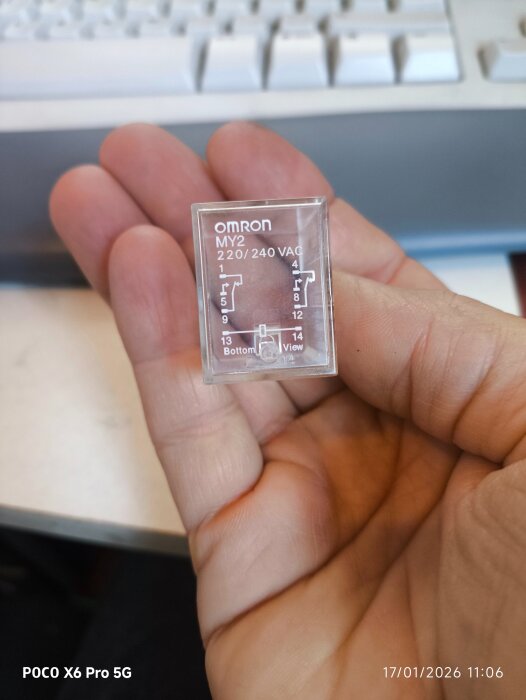 En hand håller ett transparent Omron MY2-relä med 220/240 VAC specifikationer framför ett tangentbord.