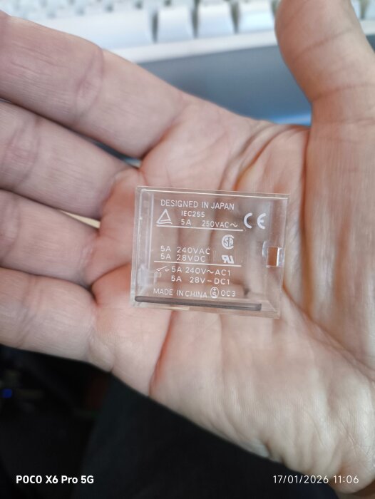 Hand som håller en transparent brytare, märkt med specifikationer 5A 250VAC. Designed in Japan, made in China. Bakgrund av dator och tangentbord.