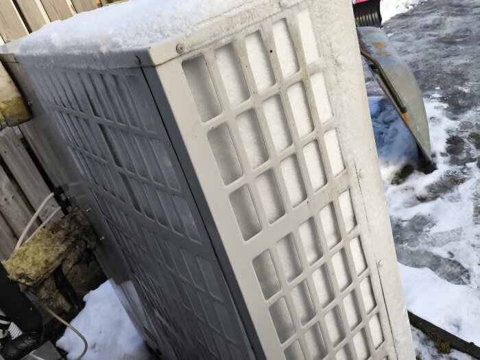 Luftvärmepump täckt med frost och snö, placerad utomhus nära ett trästaket under vintertid.
