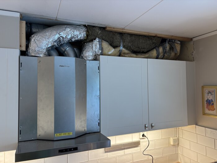 Köksfläkt med avlägsnad skåpdörr visar isolerade ventilationsrör. Under fläkten är vita kakel och en väggkontakt.
