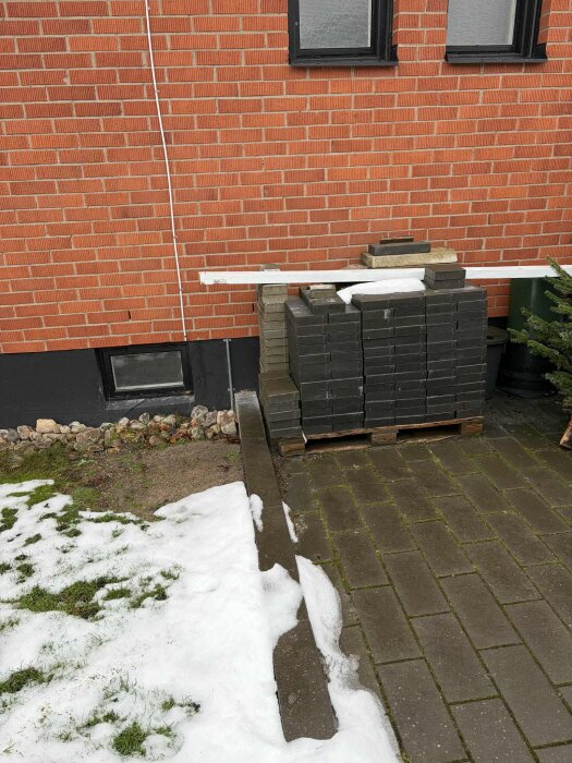 Staplade plattor vid en tegelvägg, delvis snötäckt mark och gång.
