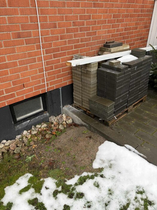 Staplade tegelstenar mot en röd tegelvägg, grus under, lite kvarvarande snö på marken.