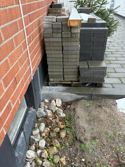 Utvändigt bygge med staplade betongplattor mot tegelvägg, synlig mark med stenar och växtlighet, samt en smältande snöhög.