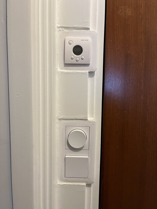 Väggmonterad vriddimmer och termostat på vit vägg bredvid dörr.