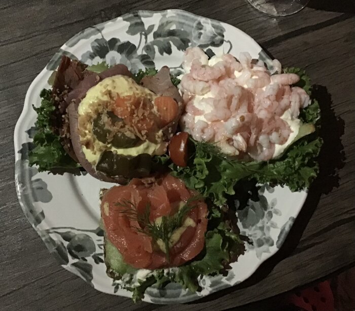 Tallrik med tre smörrebröd: ett med räkor, ett med rostbiff och grönsaker, samt ett med lax och dill, serverade på salladsblad.