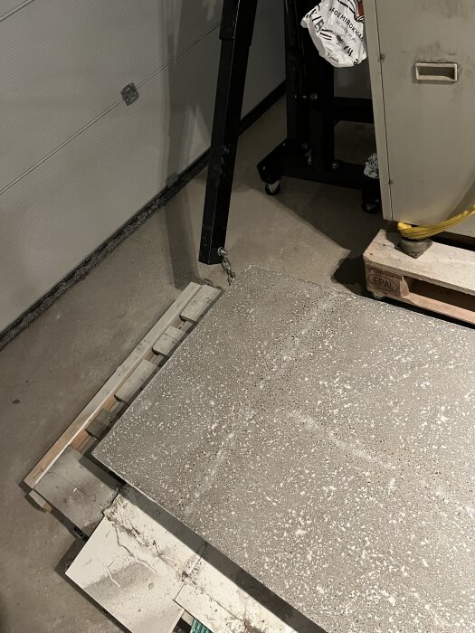 Betongplattor staplade i ett garage på en lastpall, bredvid ett stort verktyg och en säck byggmaterial.