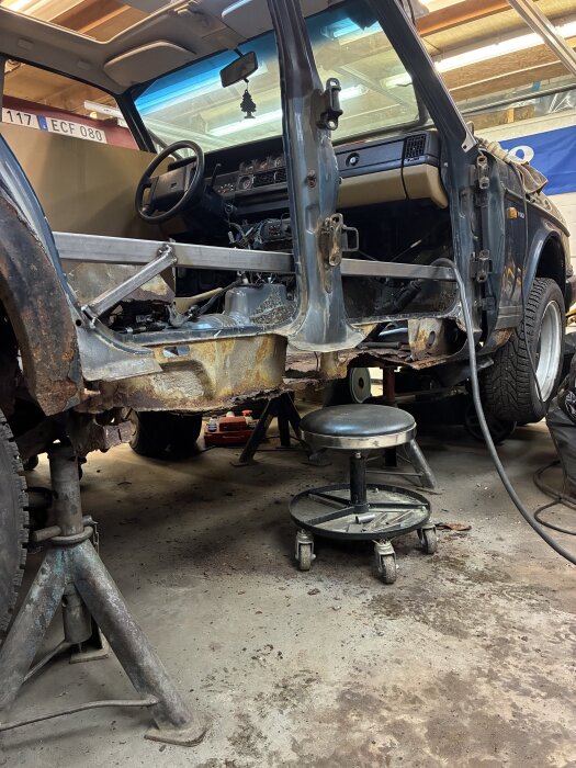En rostig Volvo 240 under renovering i ett garage, upphissad på pallbockar med förar- och passagerarsidan borttagen.