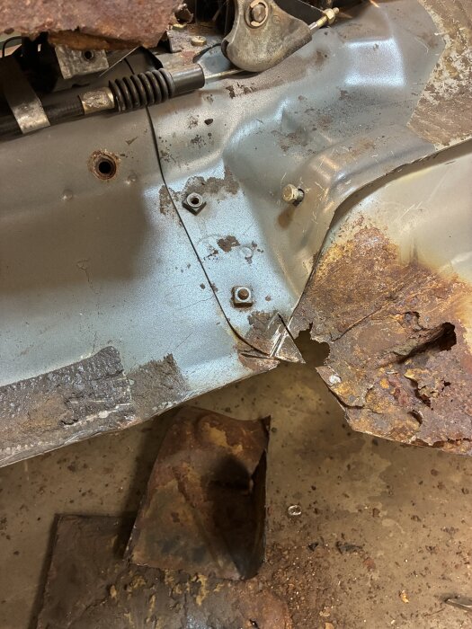 Rostig metallbit med hål och skador, del av stödlager i ett fordons chassi som exponeras i en pågående reparation eller renovering.
