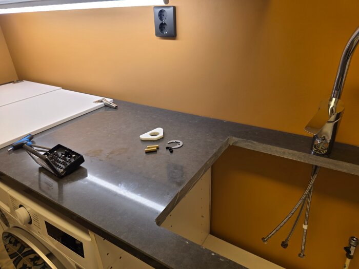 Köksbänk med verktyg, diskhoinstallation pågår. Orange vägg, vit tvättmaskin under bänkskivan, omgiven av diverse monteringstillbehör.