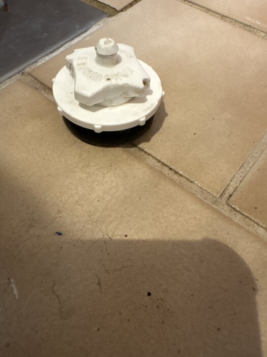 Vit plastdel med fyrkantigt fäste på kakelgolv, svårt att greppa och snurra ner.