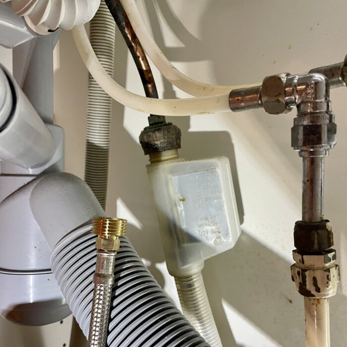 Rör och anslutningar under diskbänk i kök med kopplingar för blandare och diskmaskin. Silverfärgad och vit plastslang visas tillsammans med T-koppling.