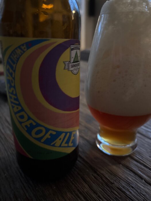 Flaska och glas med skummande ale, etikett med färgglatt mönster synlig.