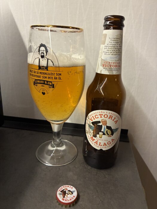 ```alt-text
Flaska och glas med Victoria Málaga öl på ett bord, med en kapsyl bredvid.
```