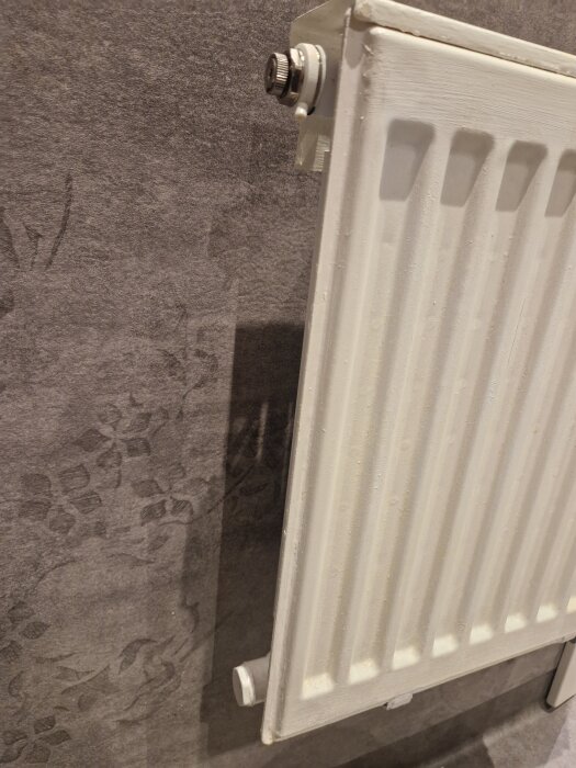 Vit väggmonterad radiator mot en mönstrad grå vägg.