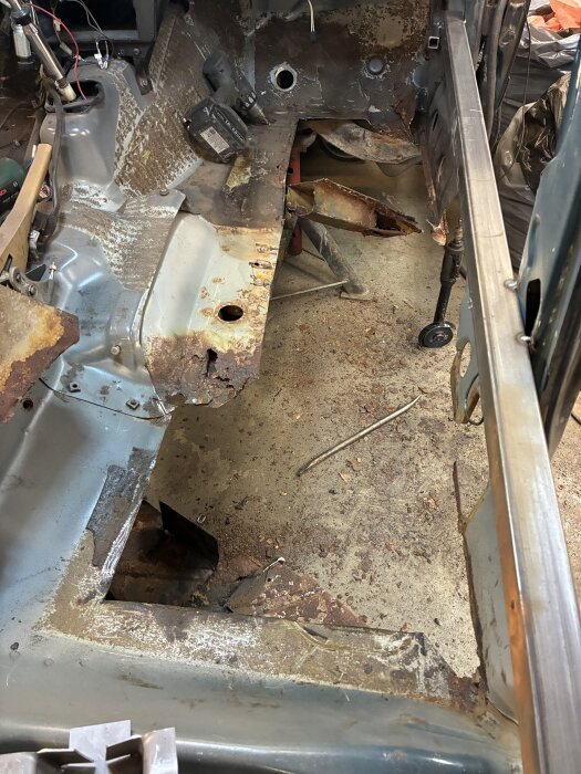 Rostskadad bilkaross med urklippta sektioner och borttagna tvärbalkar, ligger i garage med verktyg runtom.