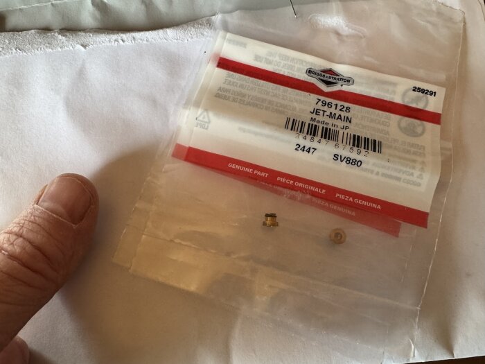 Förpackning med Briggs & Stratton jet-huvud, artikelnummer 796128, visas på en vit bakgrund bredvid en hand.