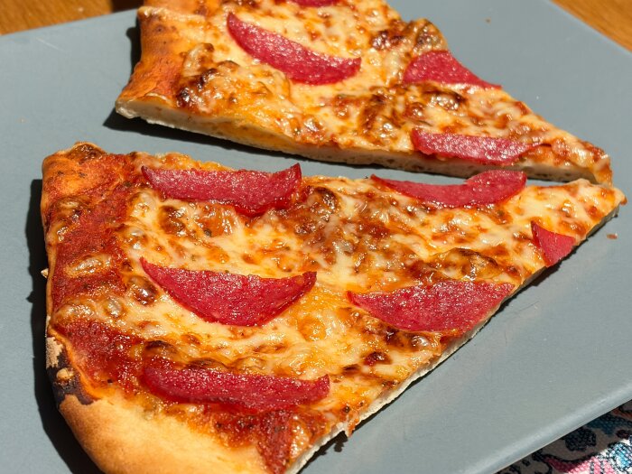 Två pizzabitar med smält ost och salamiskivor på en grå tallrik.