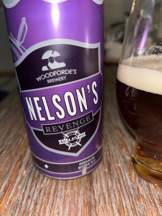 En burk Nelson's Revenge Premium Bitter Ale från Woodforde's bryggeri bredvid ett glas med öl på ett träbord.