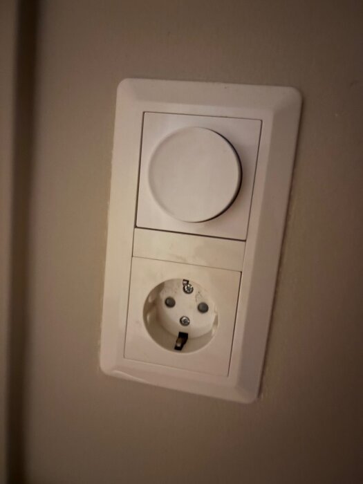 Väggmonterad dimmerknapp ovanför ett vägguttag, installerad i ett rum med elektrisk belysning.