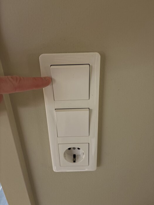 Vit strömbrytare på vägg med ett finger pekande mot den, ovanför ett vägguttag.