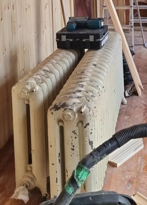 Gammal radiator med avskavd färg står lutad mot trävägg. En verktygslåda och borrmaskin ligger ovanpå.