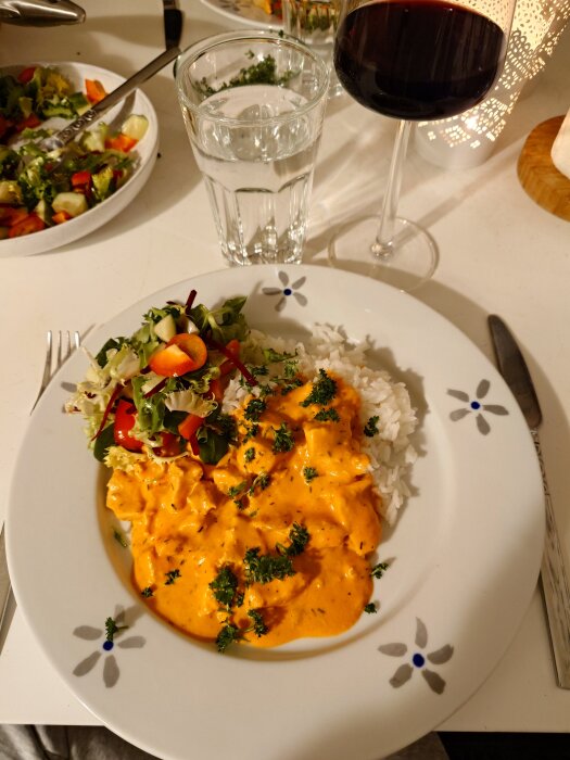 Tallrik med kyckling stroganoff och ris, serverat med sallad. Glas med vatten och rött vin i bakgrunden.