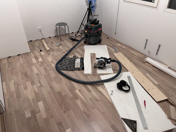 Nyinstallerat trägolv med verktyg som cirkelsåg, hörlurar och mätverktyg spridda omkring, ljusa väggar och en stol i bakgrunden.
