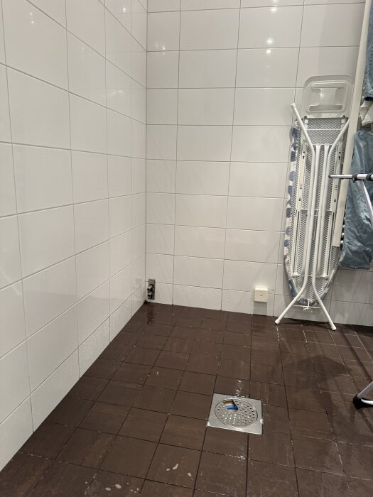 Badrumshörna med vita kakelväggar, brun kakelgolv, en elfördelningsdosa, golvbrunn och en hopfälld tvättbräda stående mot väggen.