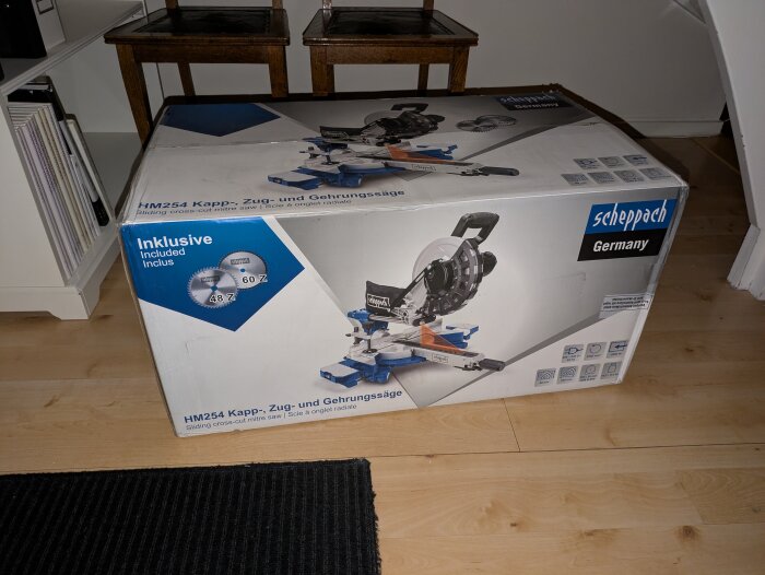 Kartong med en Scheppach kap-, drag- och geringssåg HM254 på ett trägolv, visar inkluderade sågklingor.