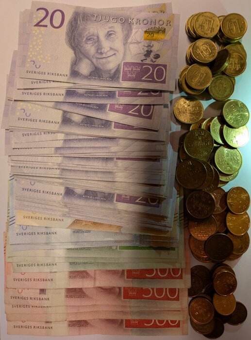 En hög av svenska sedlar i valörerna 20, 100 och 500 kronor tillsammans med en hög av svenska mynt placerade på ett bord.