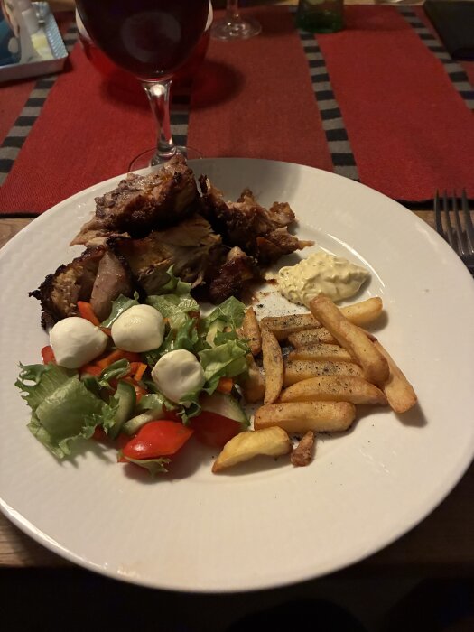 Tallrik med revbensspjäll, pommes frites, sallad med mozzarella och tomater, samt sås. Bakgrund med rödvinsglas och röd duk.