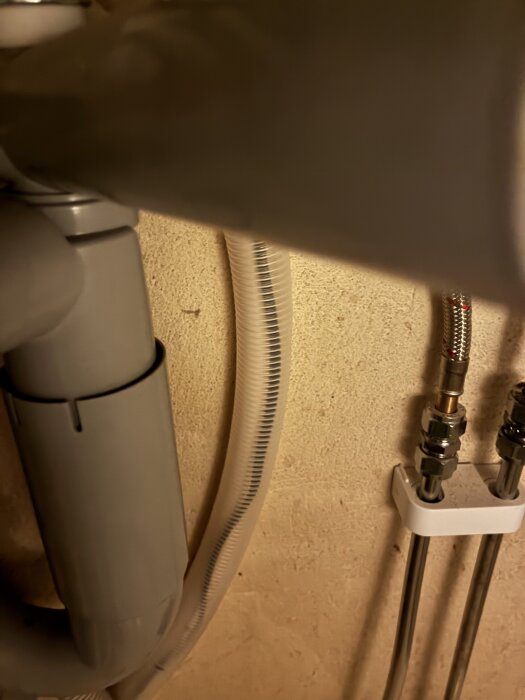 Rör och kablar i ett badrum eller kök, nära en vägganslutning, med metall- och plastdetaljer.