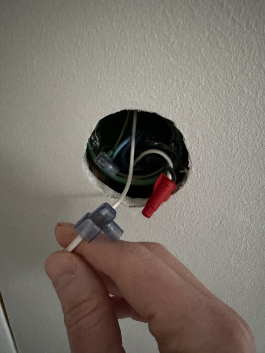 Hand håller en vit kabel framför en öppen elbox i väggen med synliga ledningar och wire connectors.