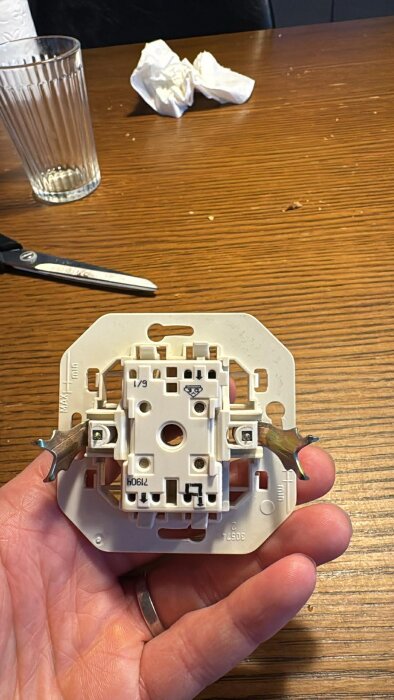 Elektrisk strömbrytare i handen, liggande på träbord med glas, papper och sax i bakgrunden.