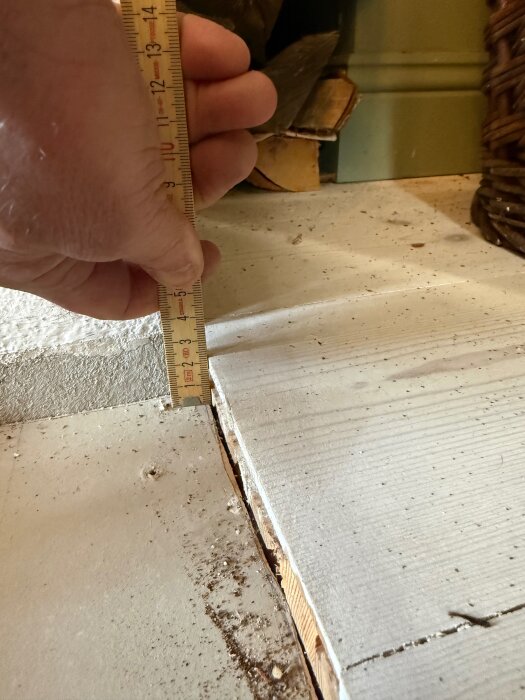 En hand håller en linjal som mäter avståndet mellan en planka och ett underlag, visande 22 mm kvarvarande planktjocklek.
