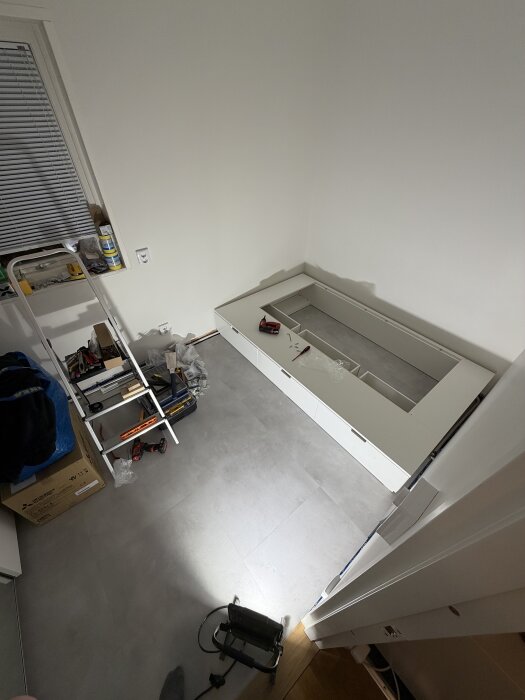 Verktyg och material för ett byggprojekt i ett barnrum, inklusive en halvmonterad sängram på ett grått klinkergolv och en stege med verktyg.